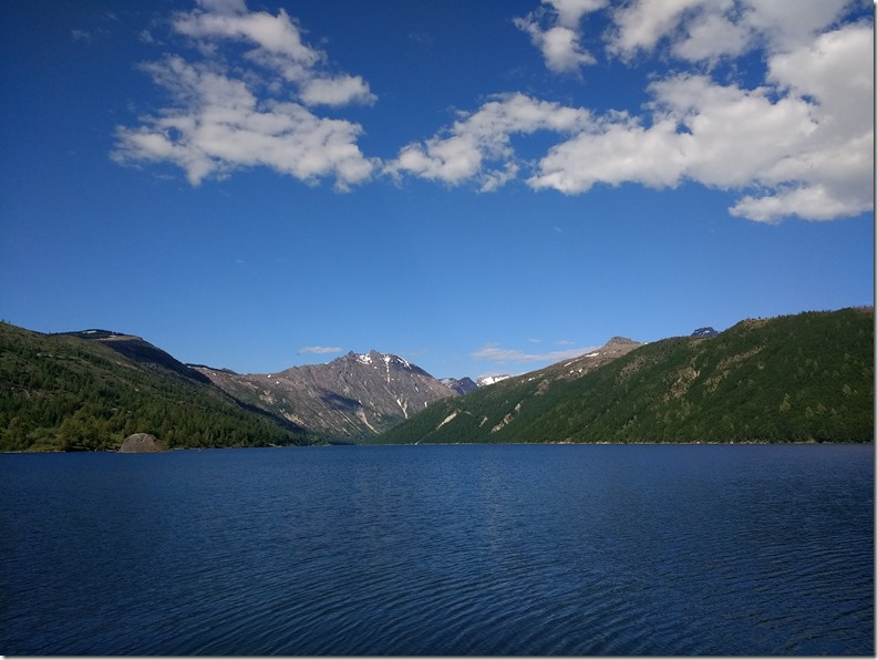 Deep blue lake surrounded by forested hills and distant mountains against a blue sky