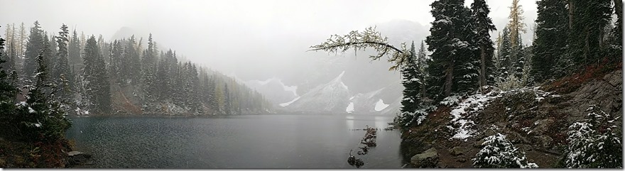 An alpine lake is surrounded by light snow on evergreen trees. It's foggy and difficult to see across the lake