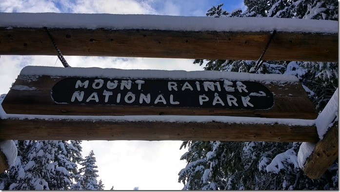 A view up at a wooden sign which says Mt Rainier National park. It's winter and the sign and surrounding trees are covered in snow