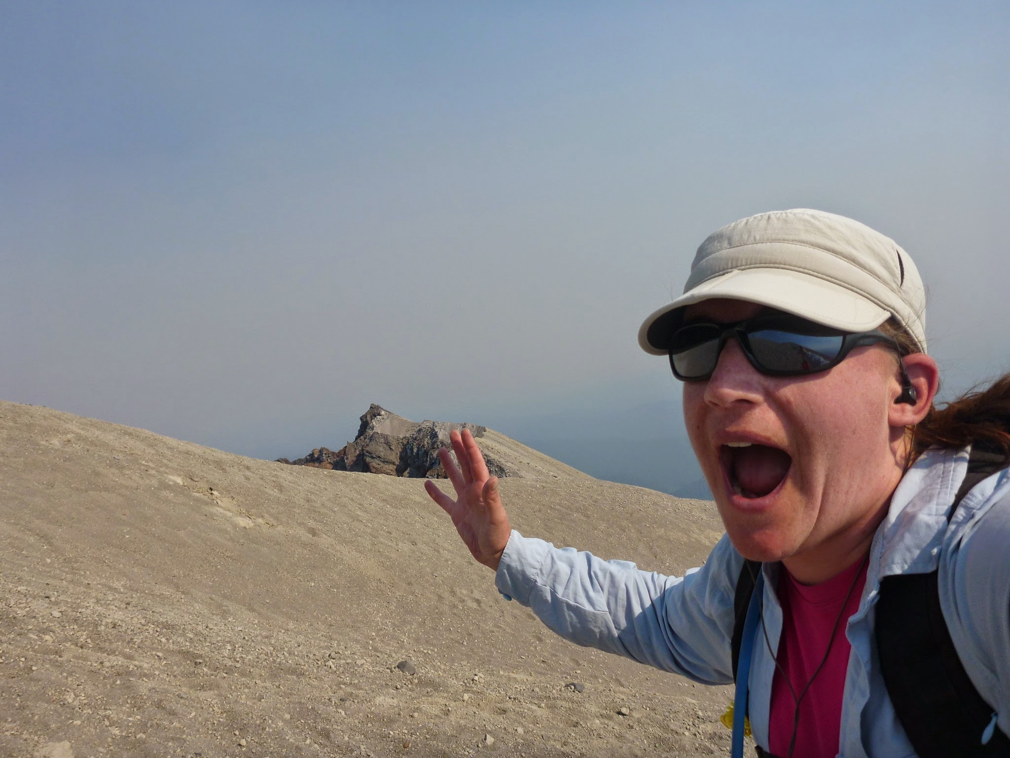 A hiker with a hand in the air, mouth open, a hat and sunglasses, near the summit of a volcano covered in sand