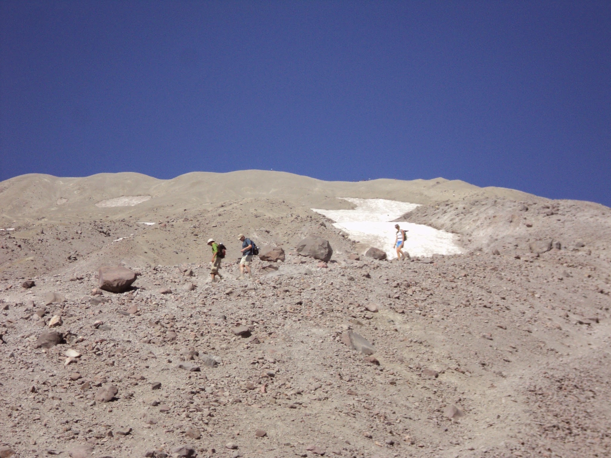 Volcanic gray sand at the top of a mountain. Three hikers walking across it. Snowfield and blue sky in the distance