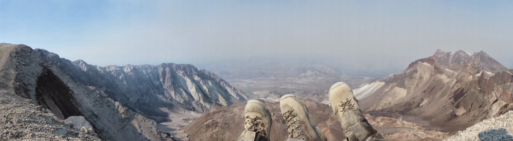 Three feet in hiking boots against a backdrop of a volcano crater, with wildfire smoke in the distance