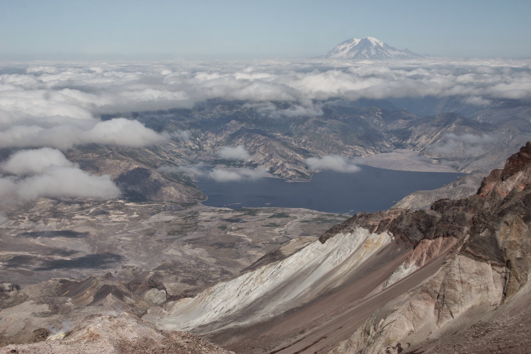 Brown volcanic crater in the foreground, a lake and hills in the distance and a high mountain in the far distance at the summit of a Mt St Helens climb