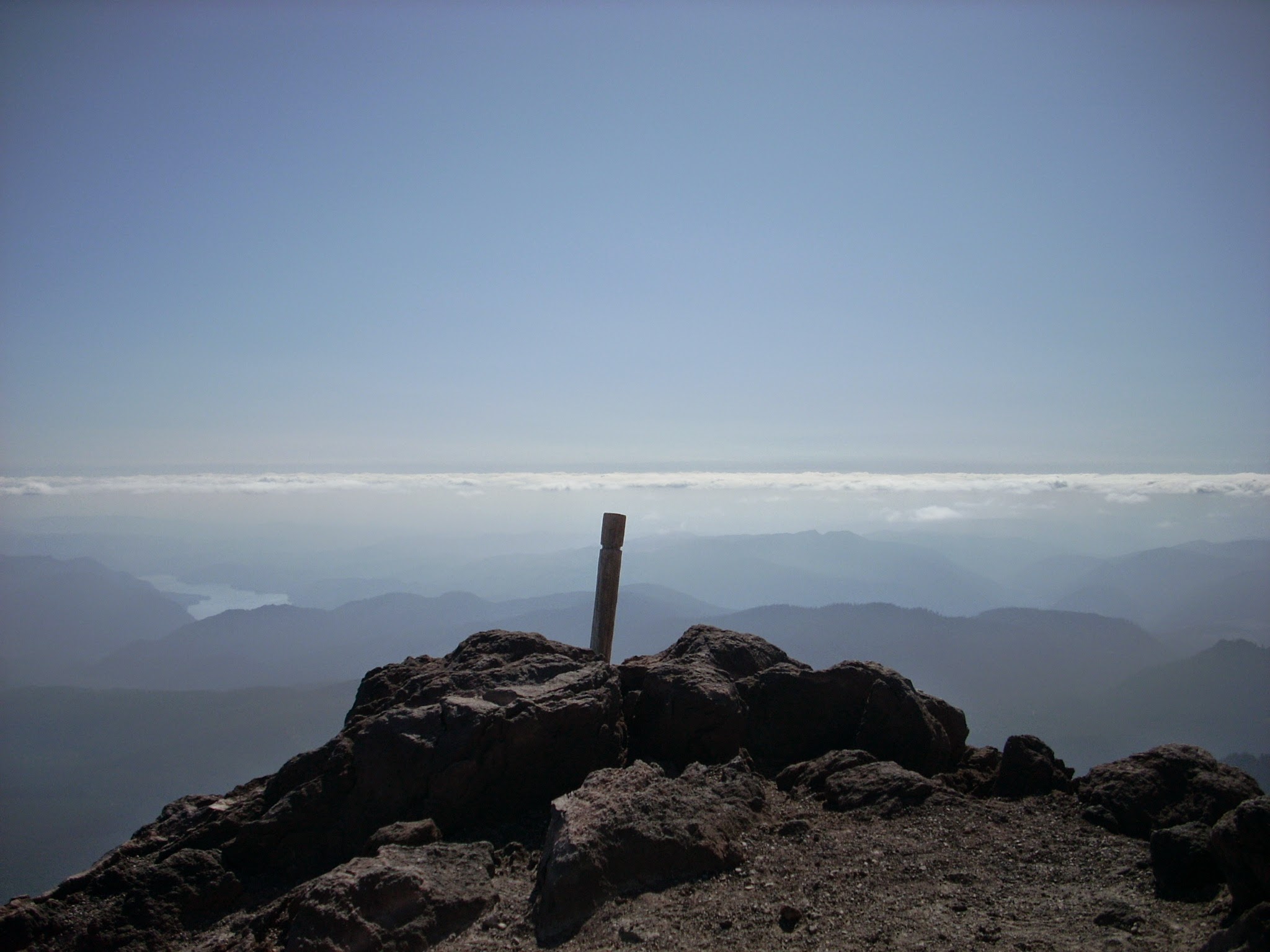 Brown rocks in the foreground with a wooden pole. Hills, a lake, clouds and blue sky in the background