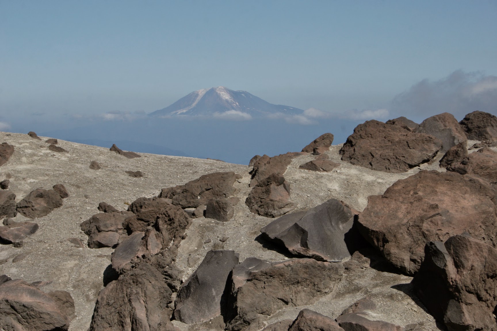 Brown rocks and volcanic sand in the foreground, a mountain and clouds in the background