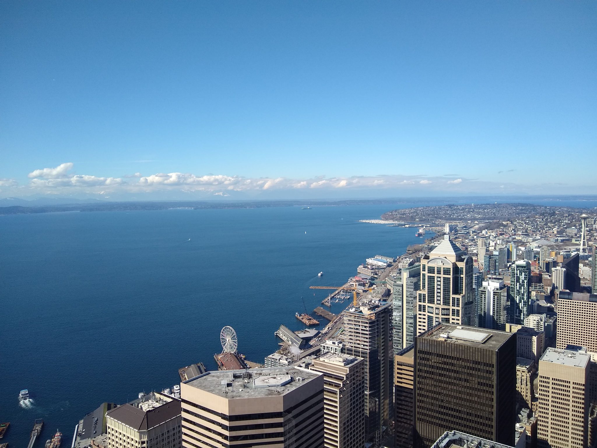 A city seen from high above. There is blue water, blue sky and some clouds above high rise buildings and a waterfront