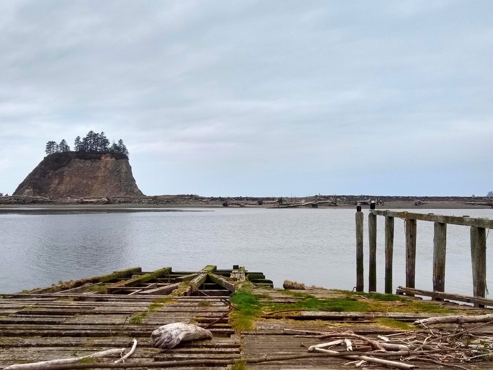 A deteriorating ramp and pier. There is a small island in the distance and a beach across a bay of the Pacific Ocean. Two eagles are on the pier