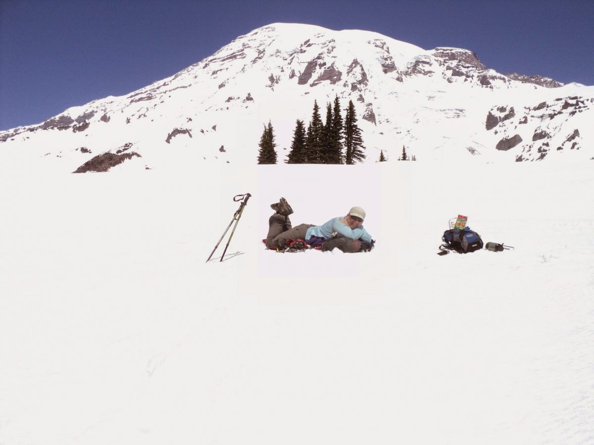 A woman laying on a backpack in a snowfield in front of a large mountain