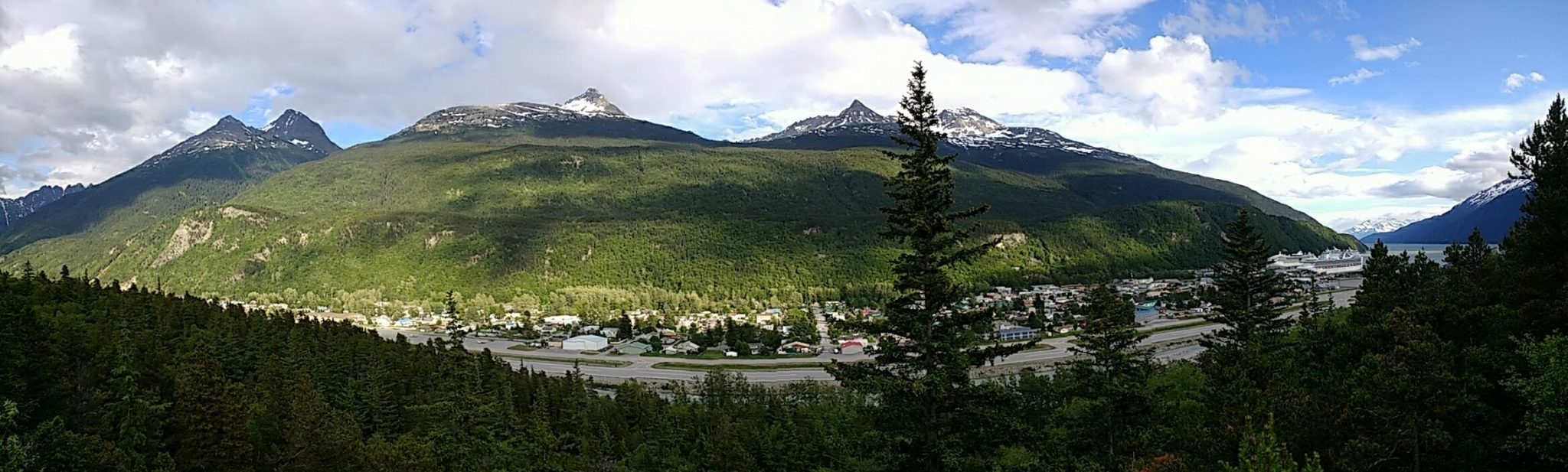 Panoramic photo of a small Alaskan town with several cruise ships in port. There are tree covered hills and snowcapped mountains in the distance. It's partly cloudy.