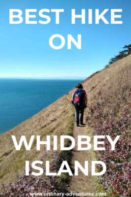 A woman is walking with her back to us on a trail on a bluff with trees to her right and blue water to her left. Distant islands are ahead of her. Text reads: Best Hike on Whidbey Island