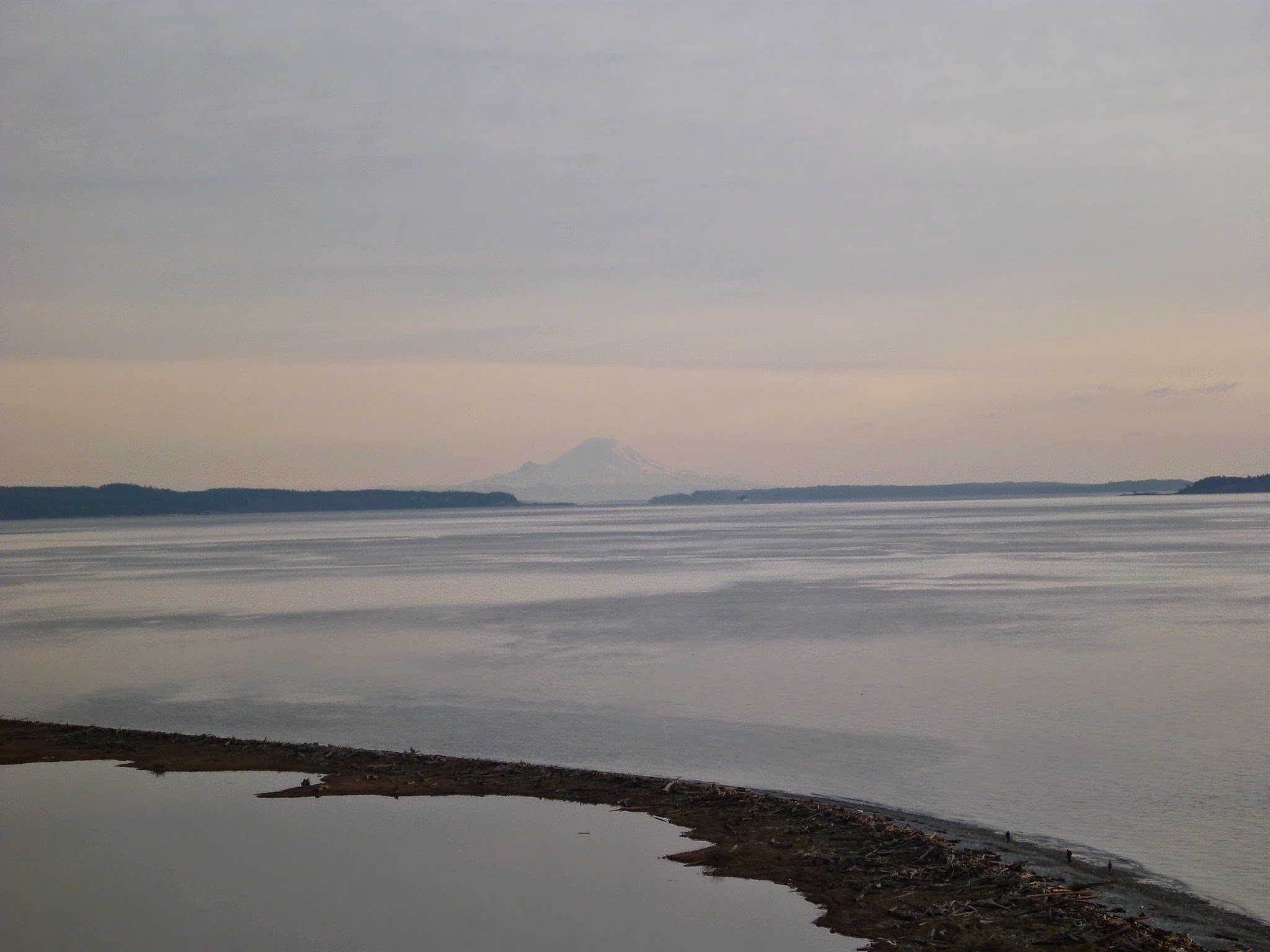 a strip of beach in the foreground at sunset on a cloudy day. Across the water there is forested land and a large snow covered mountain appears faintly in the distance