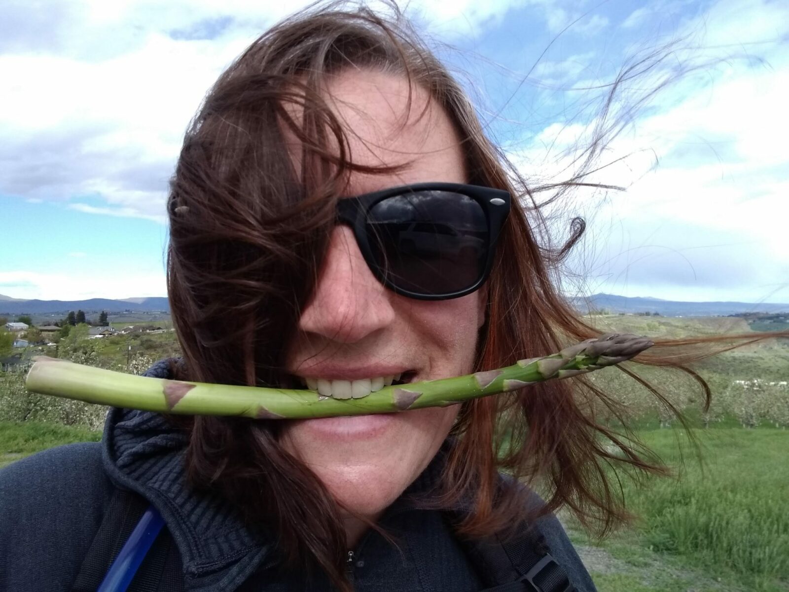 A woman wearing sunglasses is smiling and holding a single spear of asparagus in her mouth. The wind is blowing her hair from behind.