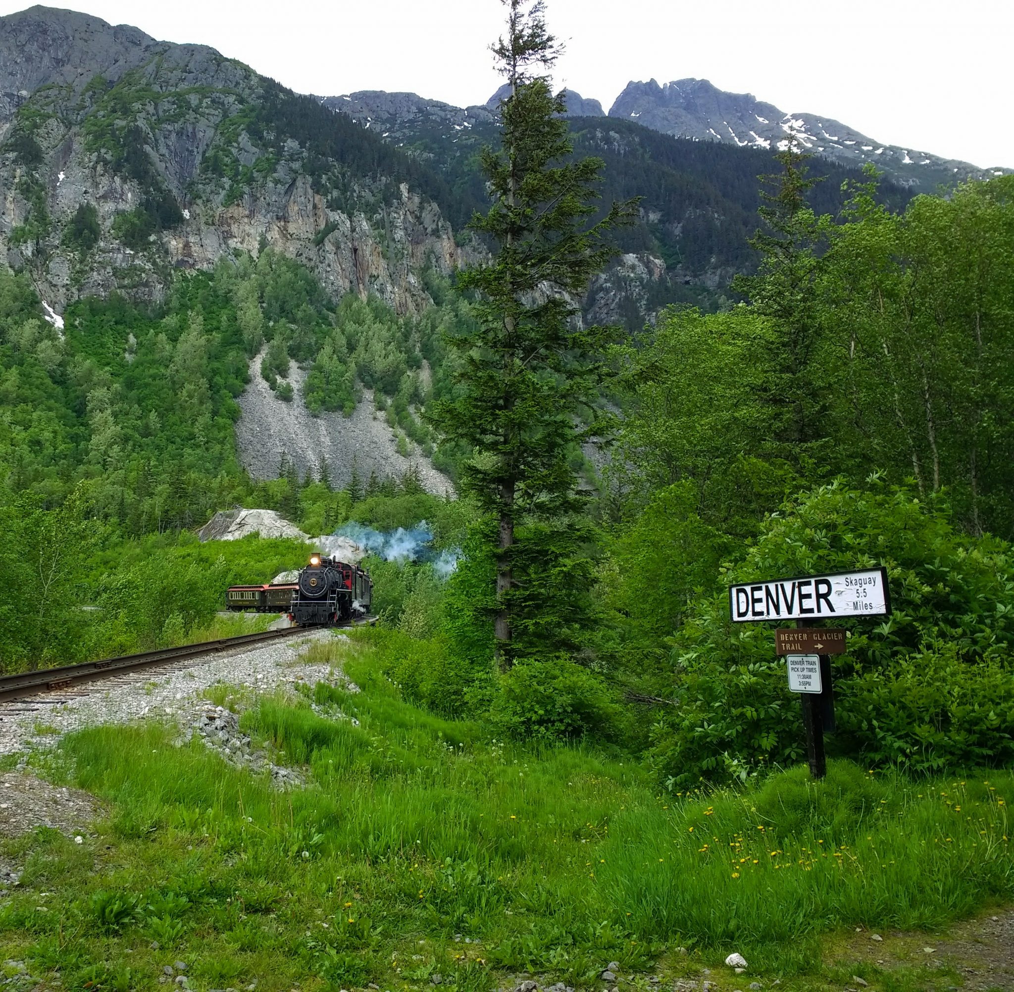 A trail approaches on a remote track, surrounded by trees and mountains. A sign says "Denver: Skagway 5.5 miles"