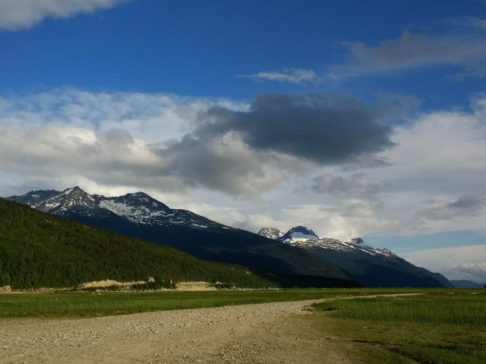 A wide flat gravel and grassy area surrounded by evergreen trees and high mountains near the Dyea campground in Alaska