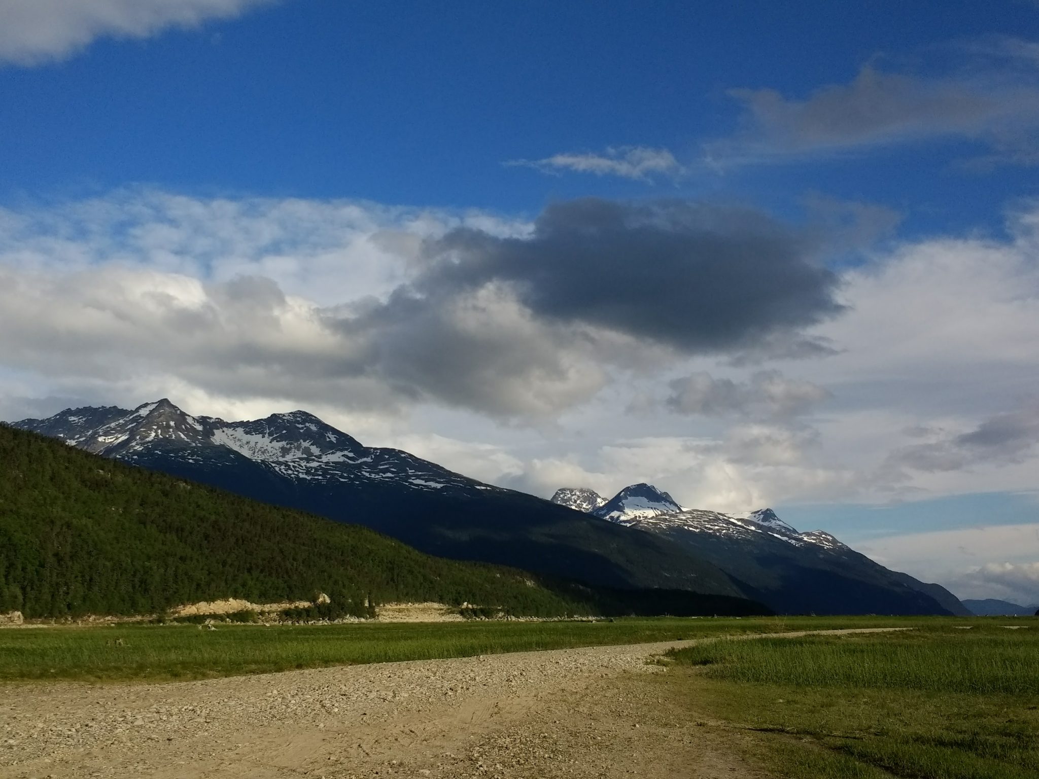 A gravel road through a meadow with snow capped mountains and partly sunny sky