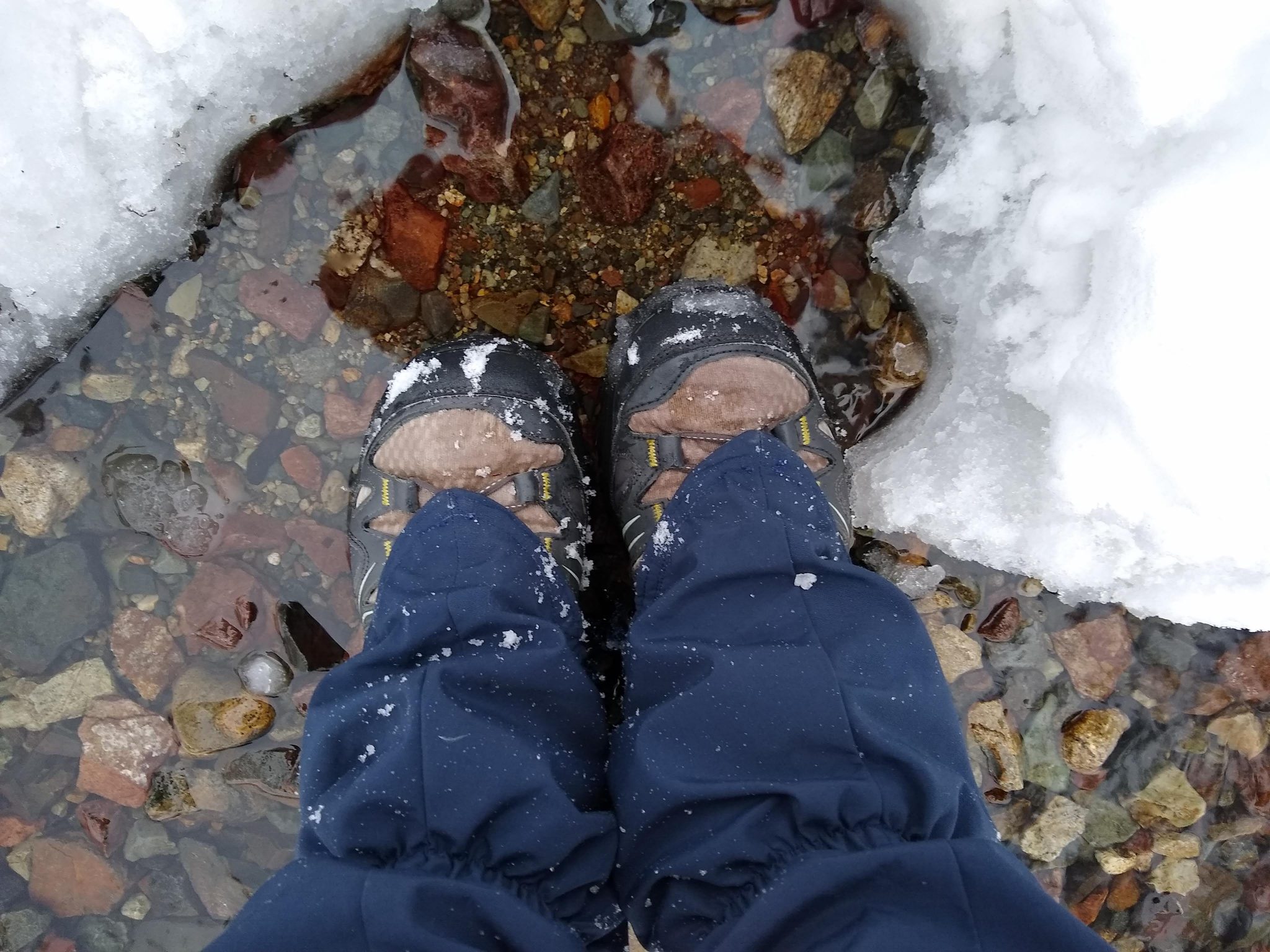 A person's lower leg and feet. They are standing in a puddle surrounded by snow. They are wearing waterproof shoes and gaiters to protect their feet and legs from getting wet