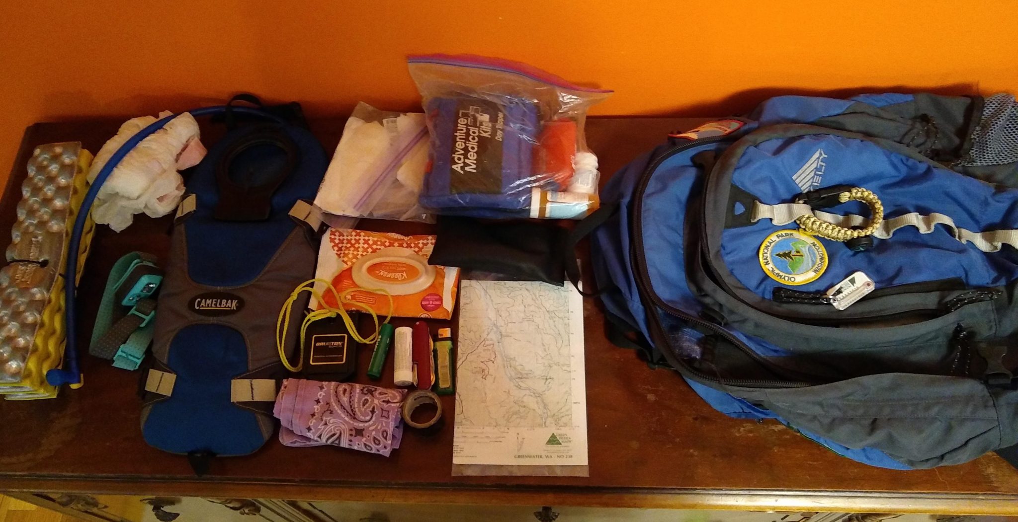 A backpack and some of it's contents spread out on a table including a map, first aid kit, water bladder, headlamp, bandana and plastic bag.