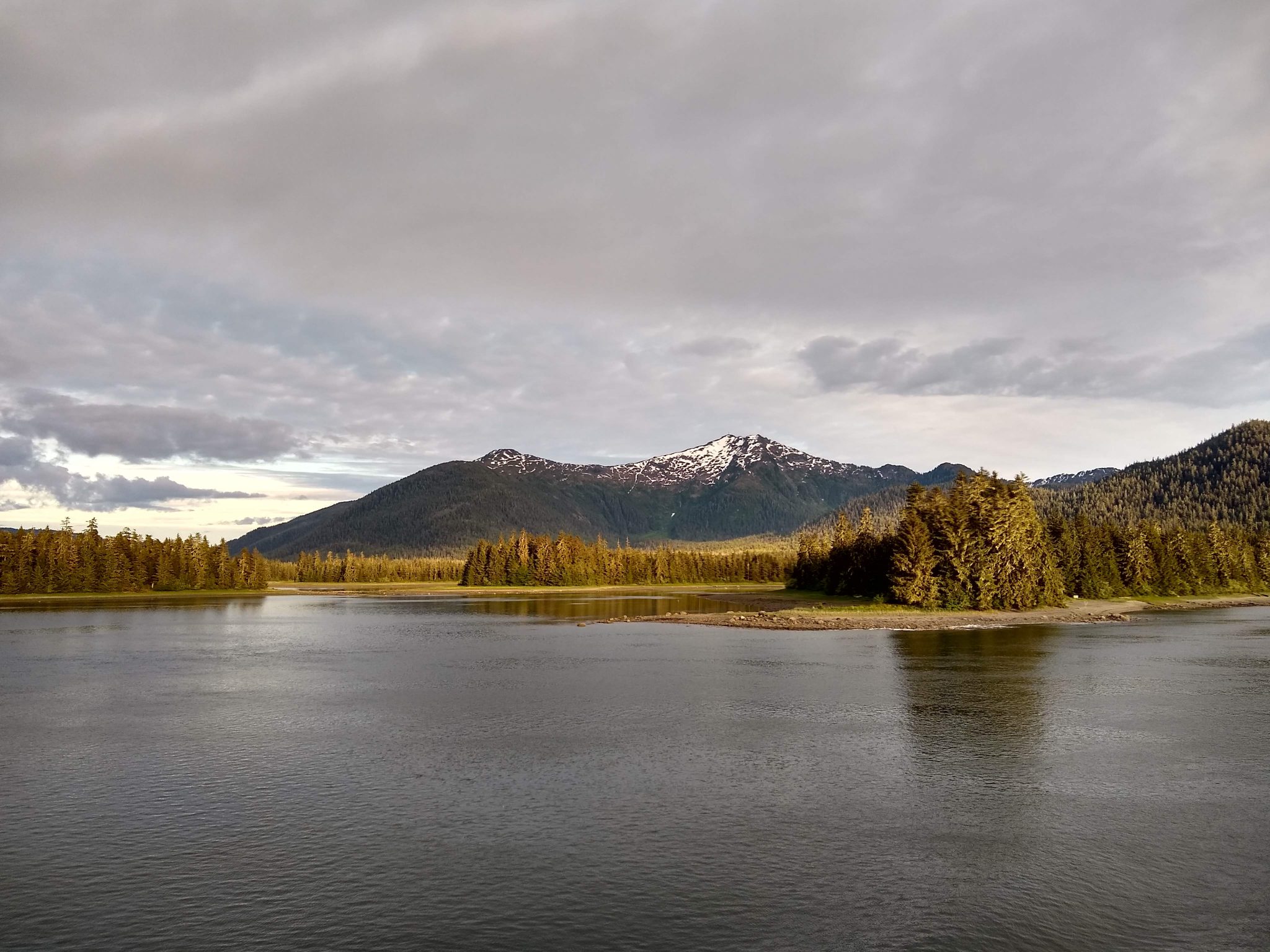 Calm waters in the foreground, green trees and higher mountains with a bit of snow in the distance on a cloudy day