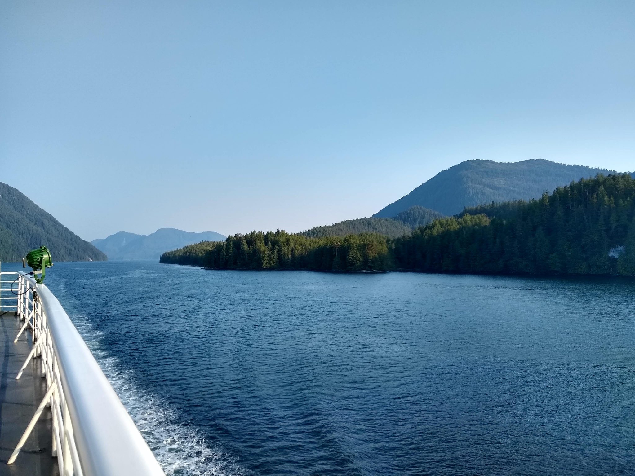 The railing and deck of the Alaska ferry are visible as it passes through a narrow channel between forests hills and mountains on a sunny day