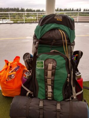 A large green backpack. It has a sleeping pad and two waterbottles attached to it, as well as a pair of sandals and a mug. An orange bag holds food.