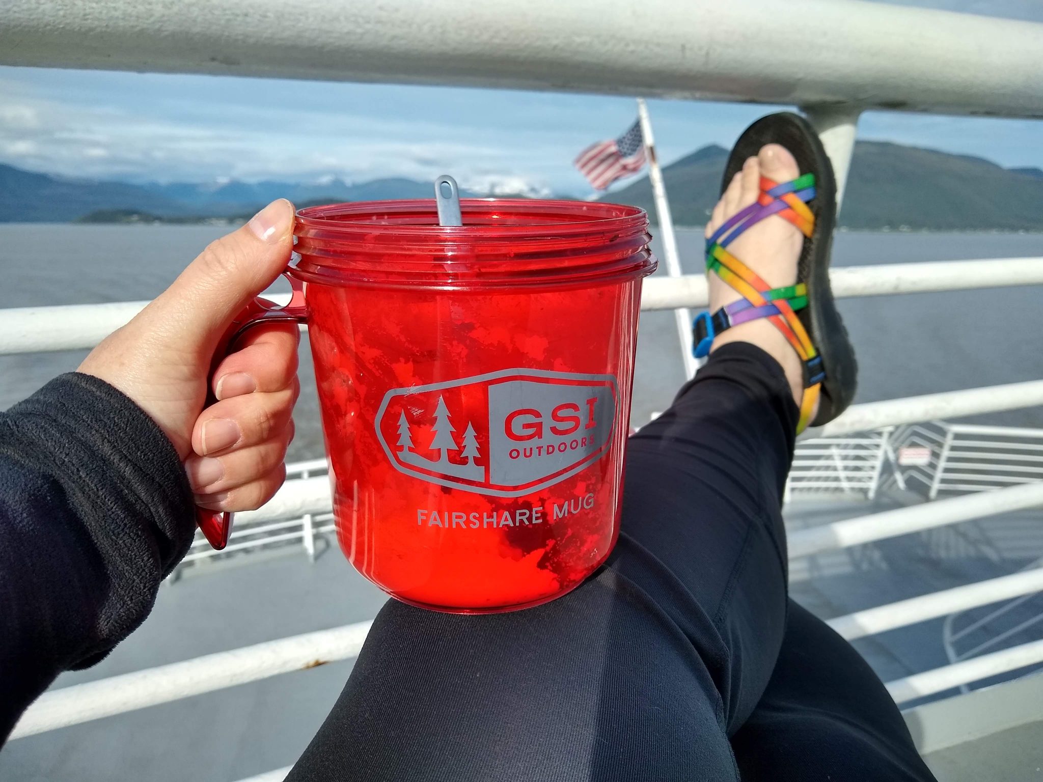 A woman is on the deck of a ship. We can see only her legs, feet and hand holding a red mug with food in it.