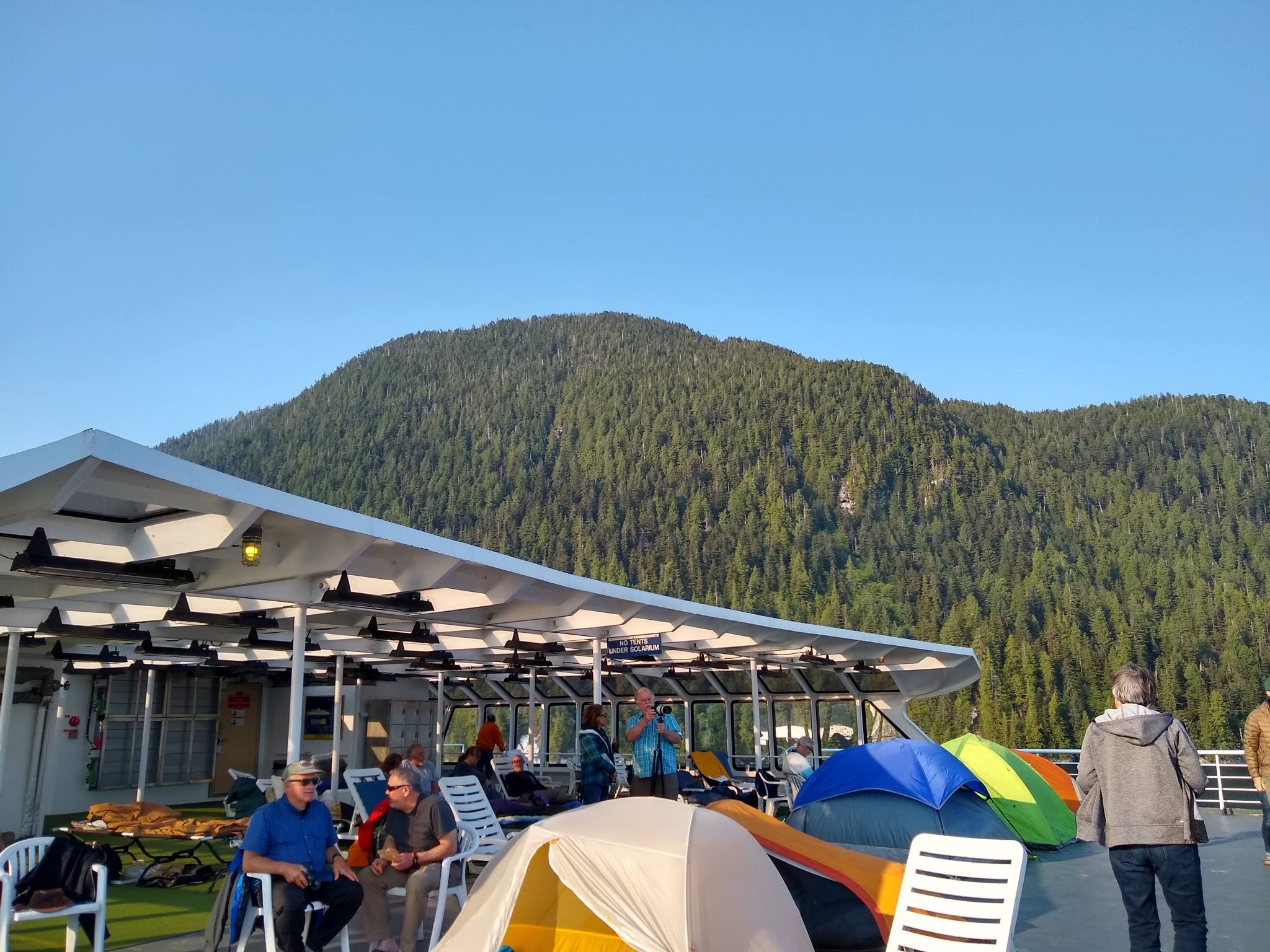 People on the top deck of the Alaska ferry. There are tents on the deck and sleeping bags under the solarium. The ferry is passing close to a forested hillside on a sunny day.