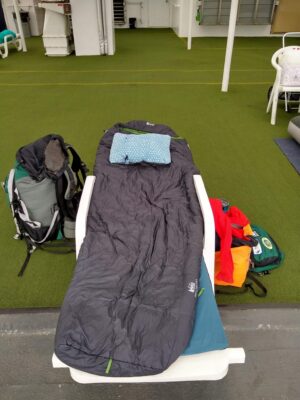 A sleeping bag, pad and pillow are on a lounge chair laying flat on the deck of the Alaska ferry. Luggage is sitting next to it.