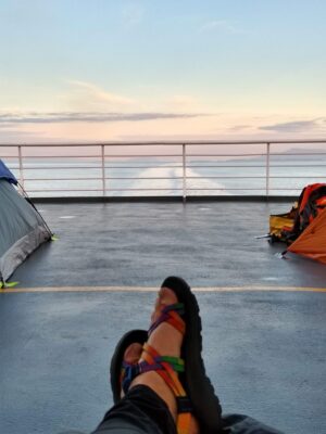 A woman's feet are visible in front of her. She is on the deck of a ship at dusk. There are two tents visible on the deck on either side of her.