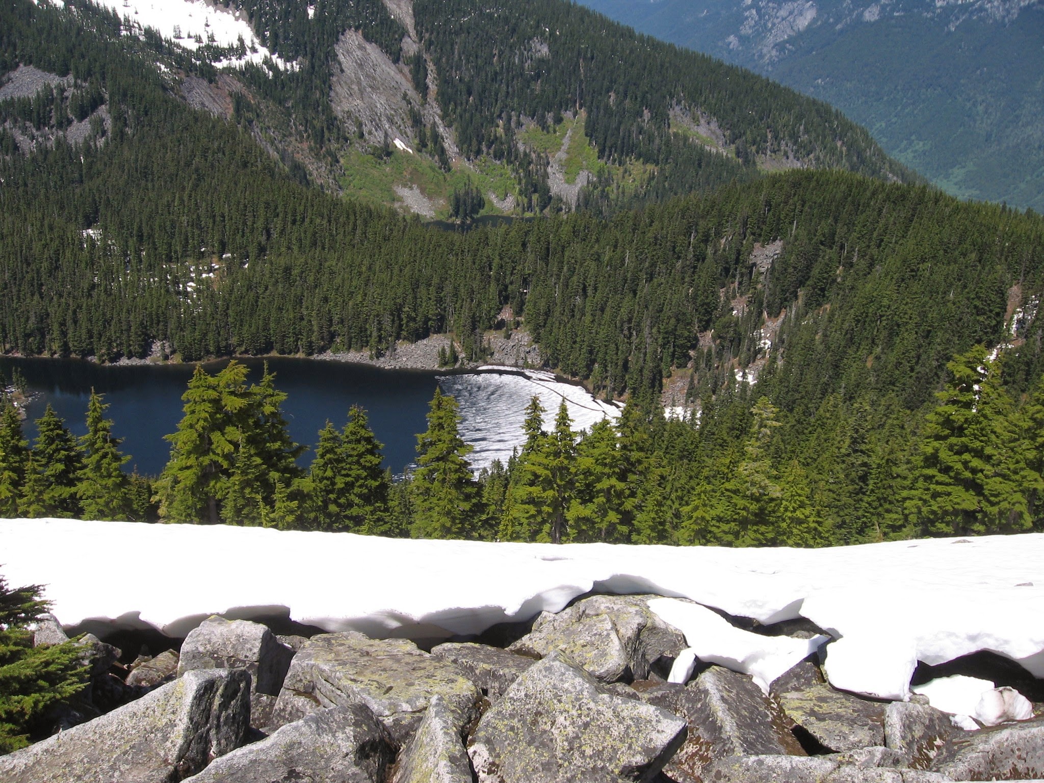 Snow covered rocks in the foreground, with trees and an alpine lake and other mountains in the background