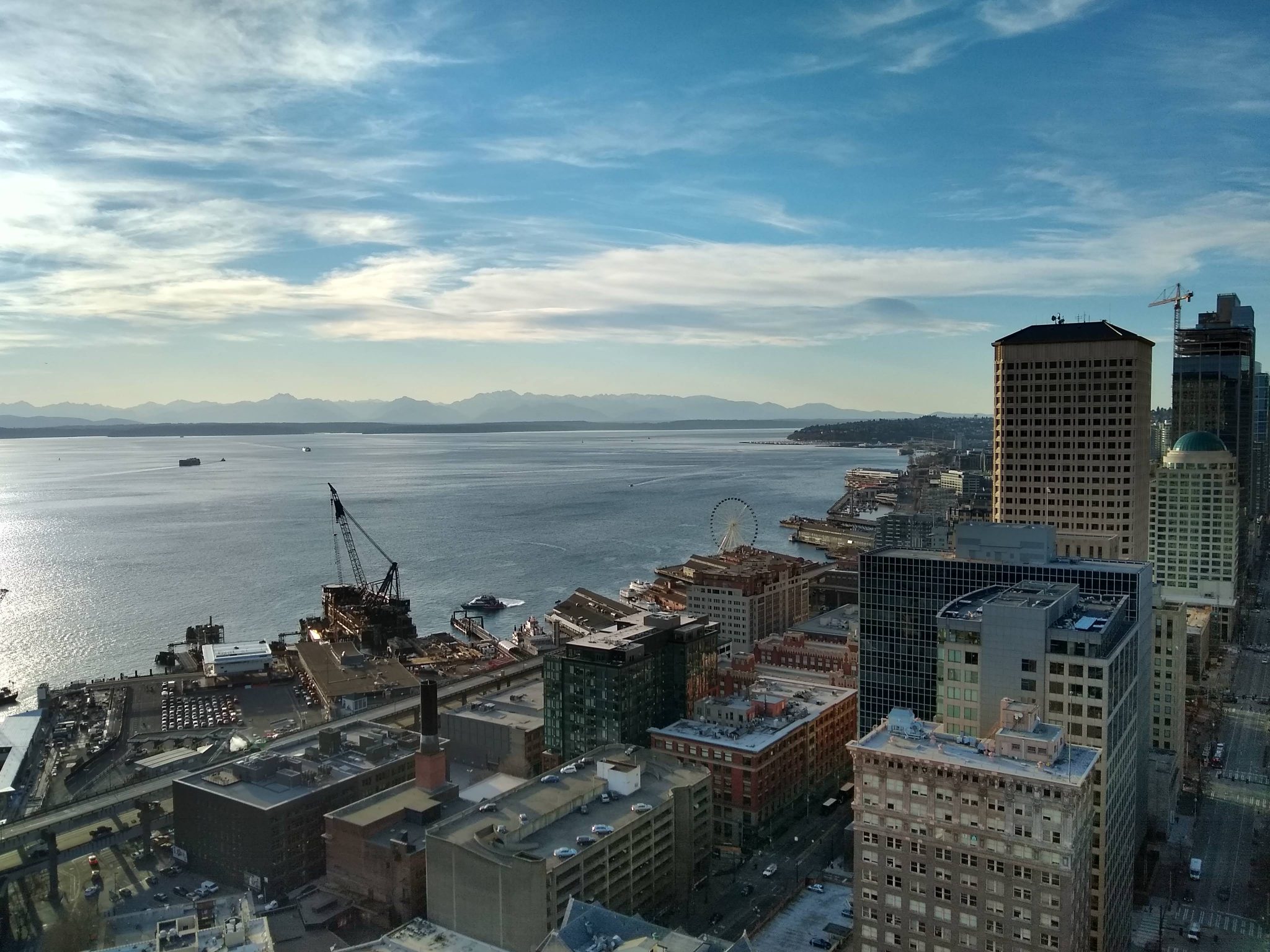 A city skyline has high buildings and a water front with distant mountains and boats on the water