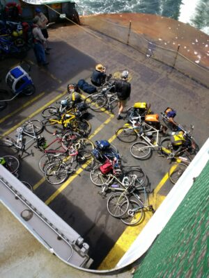A pile of bikes laying down on the deck of the ferry from above coming back from Lopez Island bike camping. A few people are sitting around the bikes.