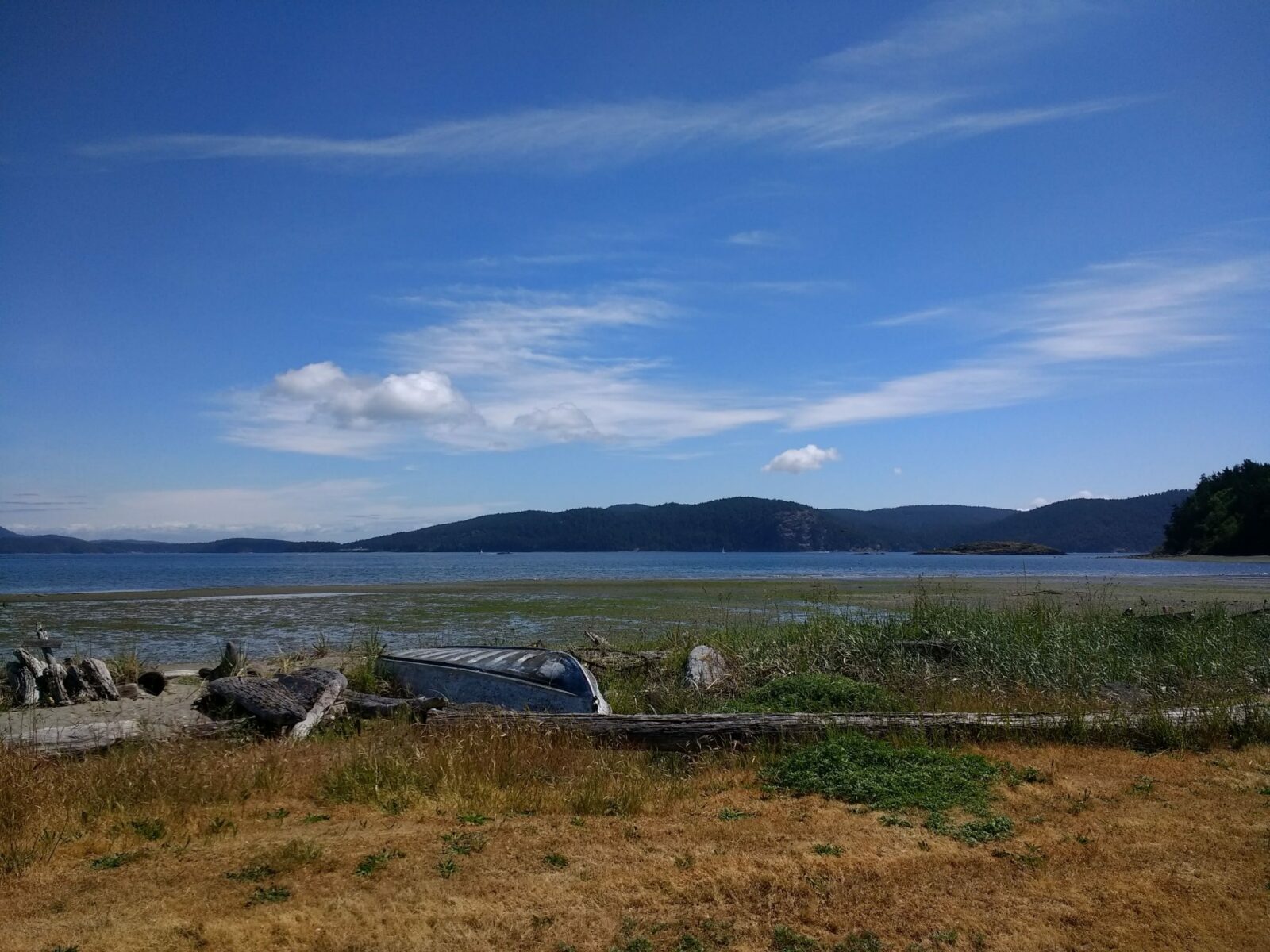 Lopez Island bike camping at Spencer Spit State Park. There is a grassy spit in the foreground wtih a canoe, tideflats and water behind. Forested islands are in the background on a sunny day