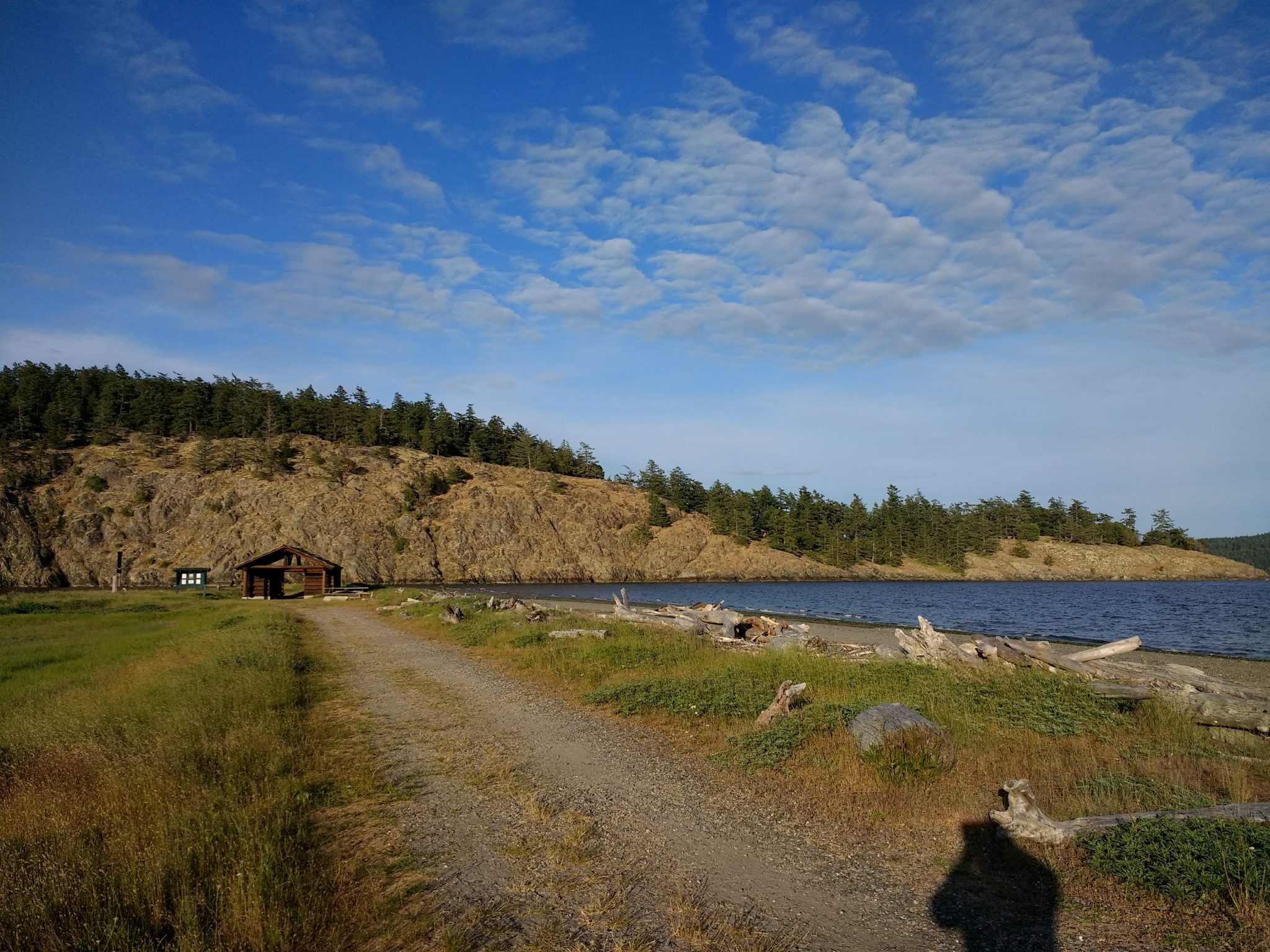 A piece of land sticks out into the water. It has a wide, gravel trail in the middle, and beach with driftwood to the side of the trail and the grass. In the distance there is a wooden shelter and a steep, forested island