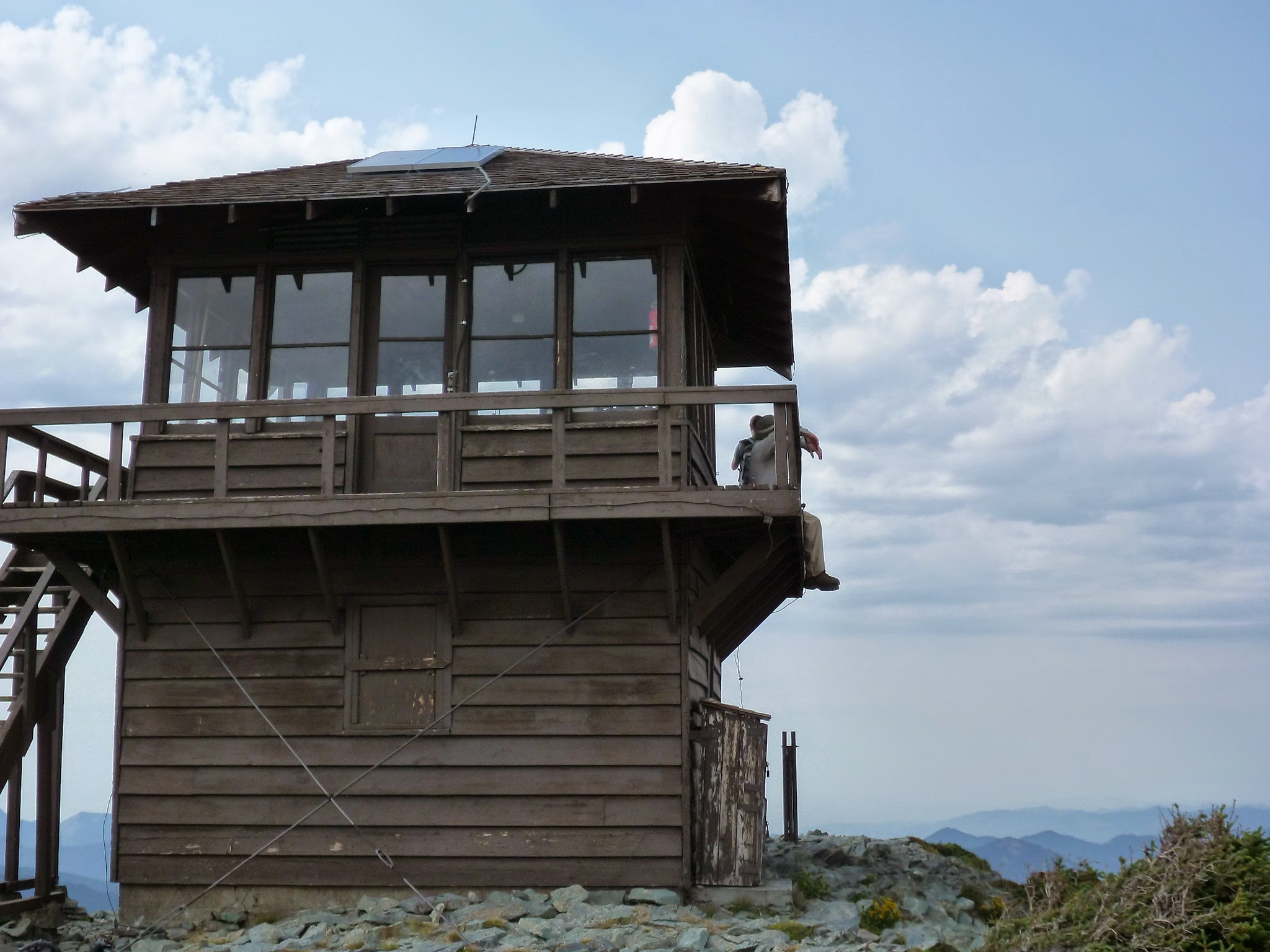 A brown wooden fire lookout tower on a pile of rocks. There are puffy clouds in a blue sky and distant mountains. Two people are sitting on the deck of the lookout tower on the Mt Fremont hike