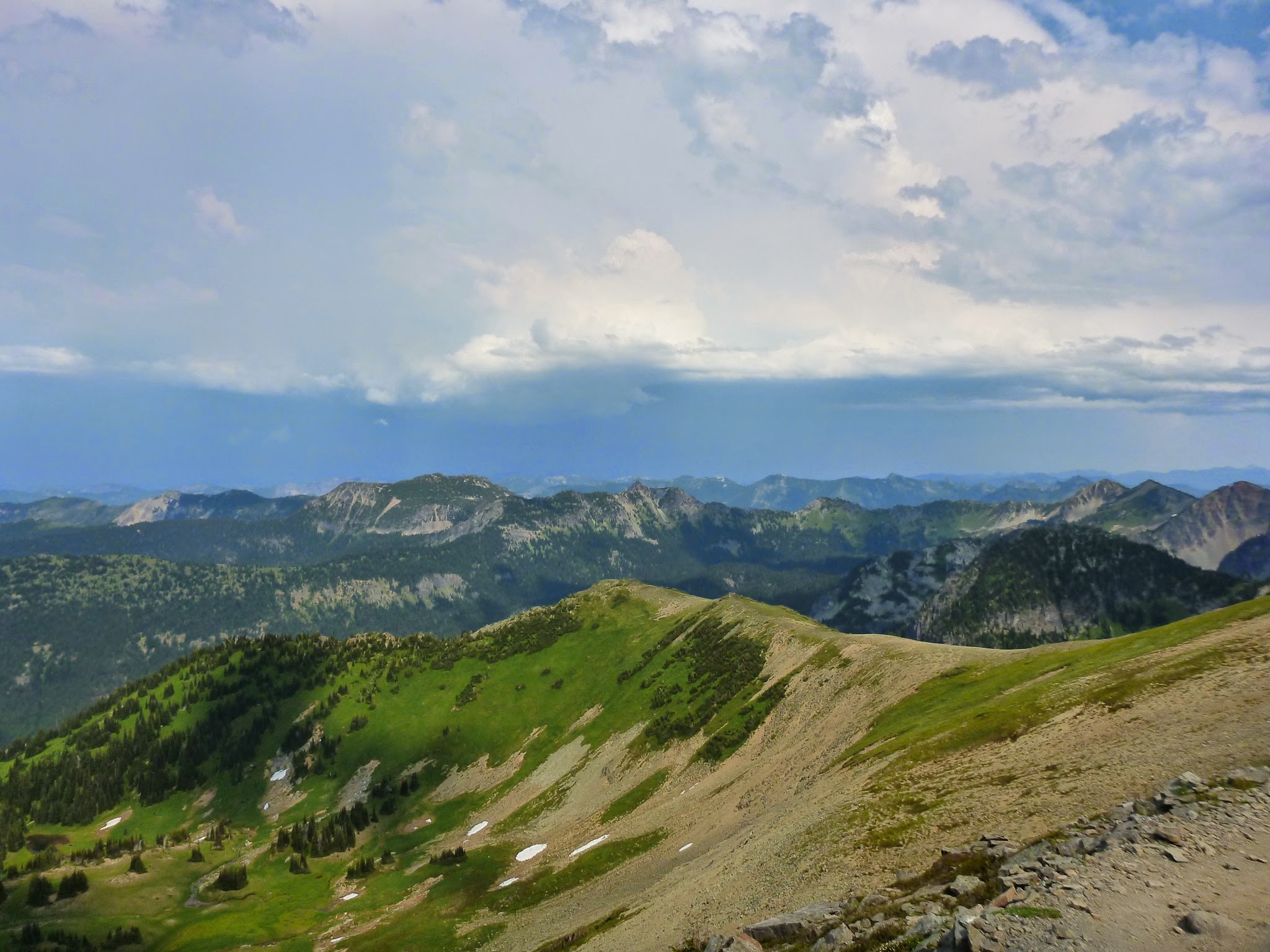 Gray clouds in the distance and distant mountains. In the foreground there's a steep green meadow with a few snow patches