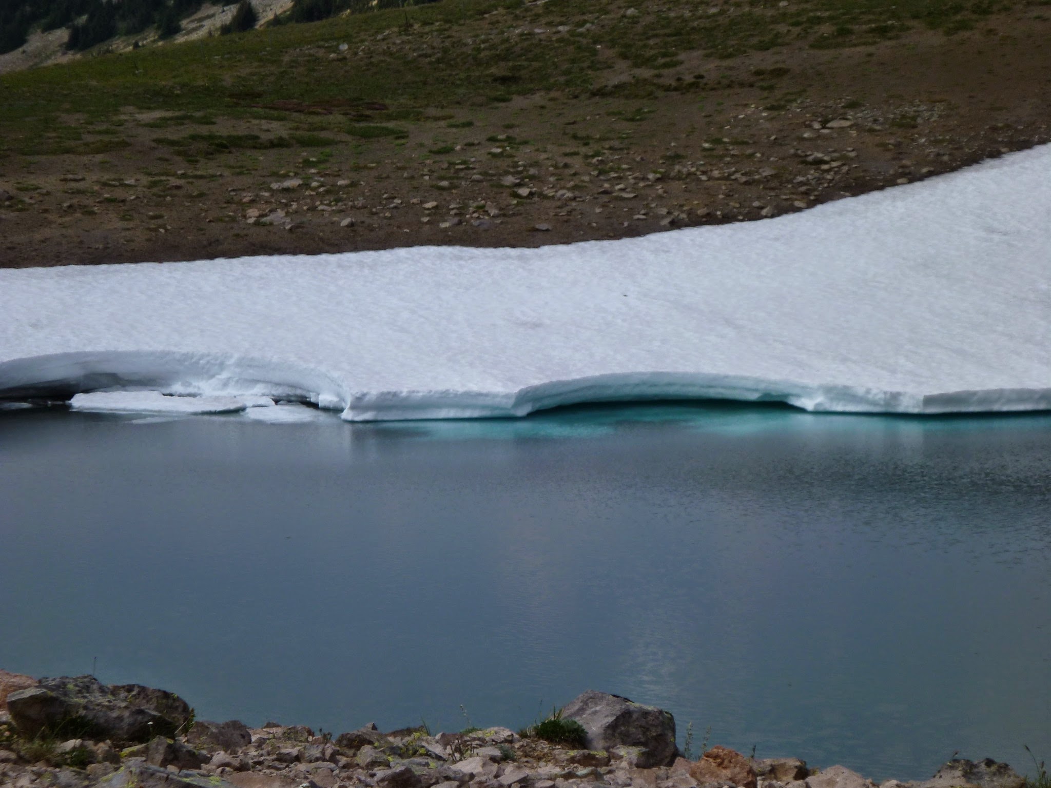 A small blue lake partially surrounded by ice and snow. Bare rocks are visible in the foreground and background