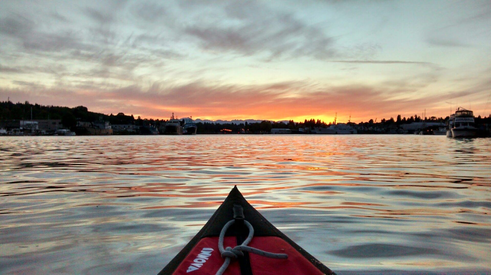 The bow of a kayak in the water just after sunset. There are boats and tree lined hills around the lake