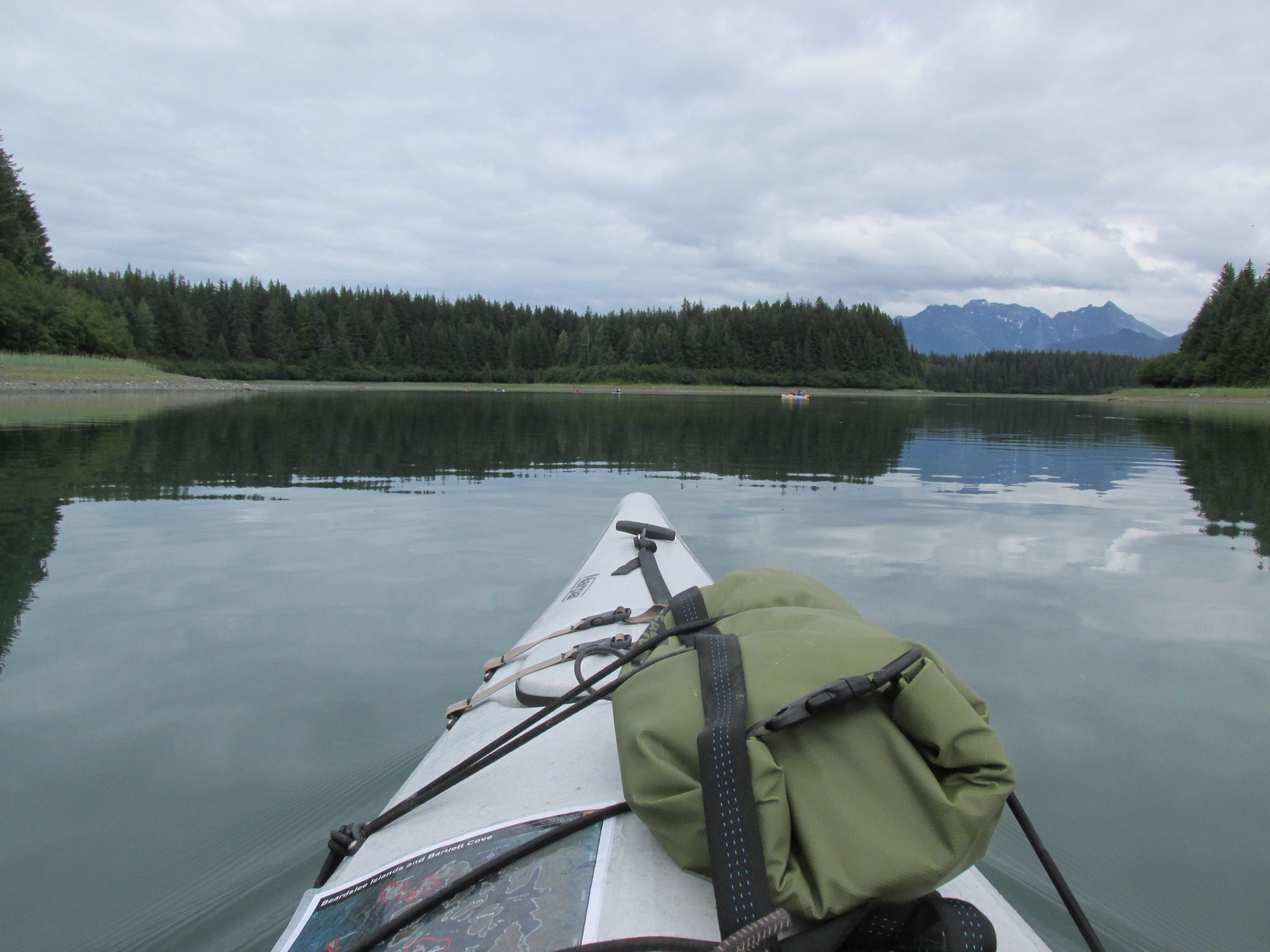 The front of a kayak with a map and dry bag tied to it. The water is calm, there are forested islands around it and some distant mountains on an overcast day