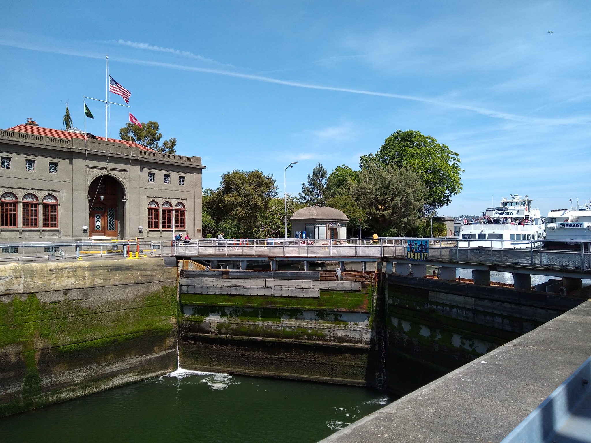 A large stone building with flags on the left. A lock with low water in front and higher water in the back with a boat in it.