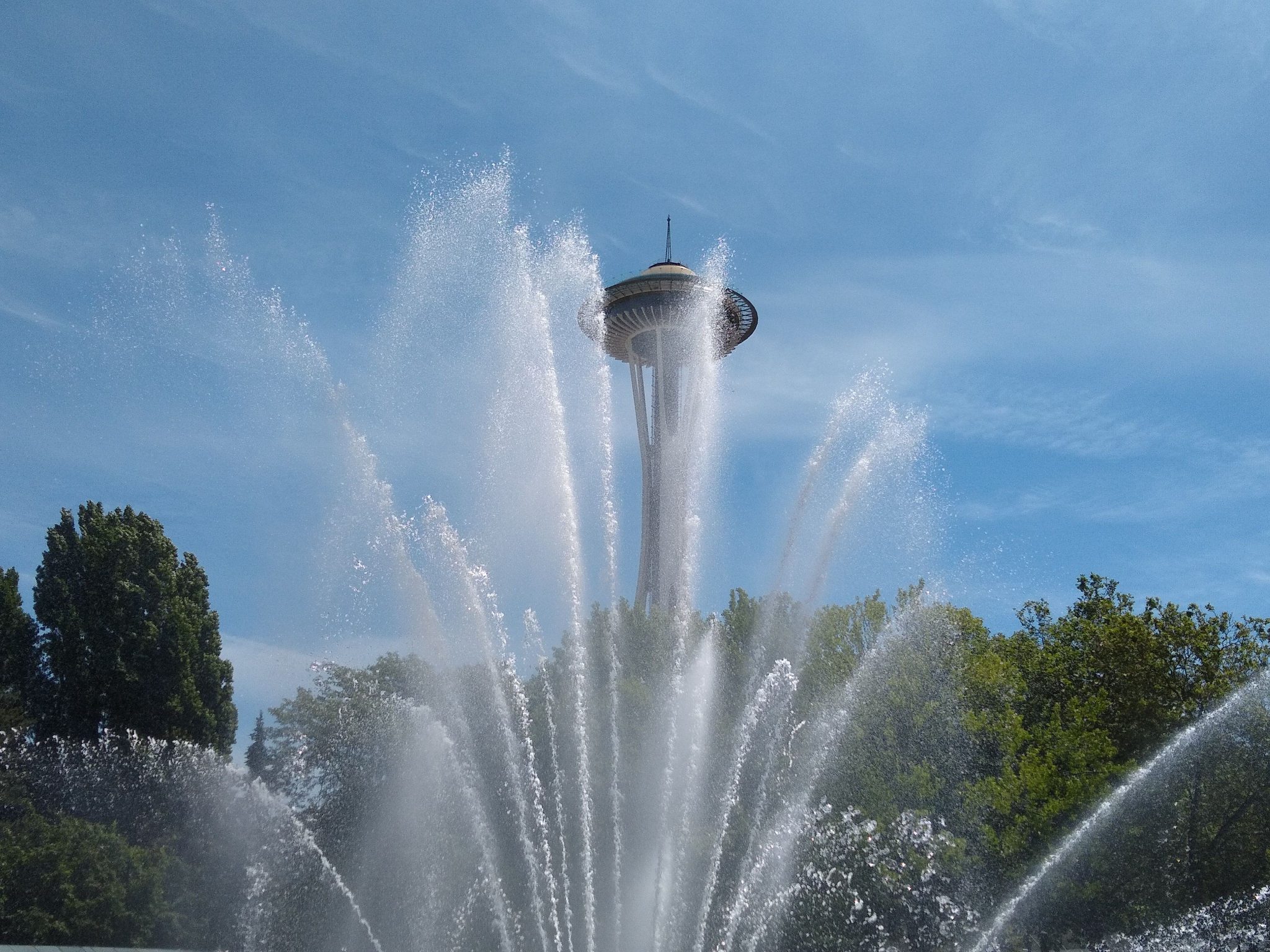 A fountain is spraying in front of green trees and the Space Needle.