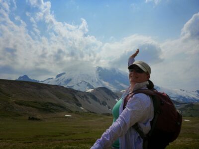 A woman is standing in front of a green meadow with clouds and Mt Rainier in the background. She is wearing sunglasses, a hat, a backpack and a long sleeved shirt. She will teach you how to start hiking!