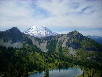 Mt Rainier behind a forested and rocky hillside which surrounds a blue, alpine lake. How to start hiking and enjoy these views!