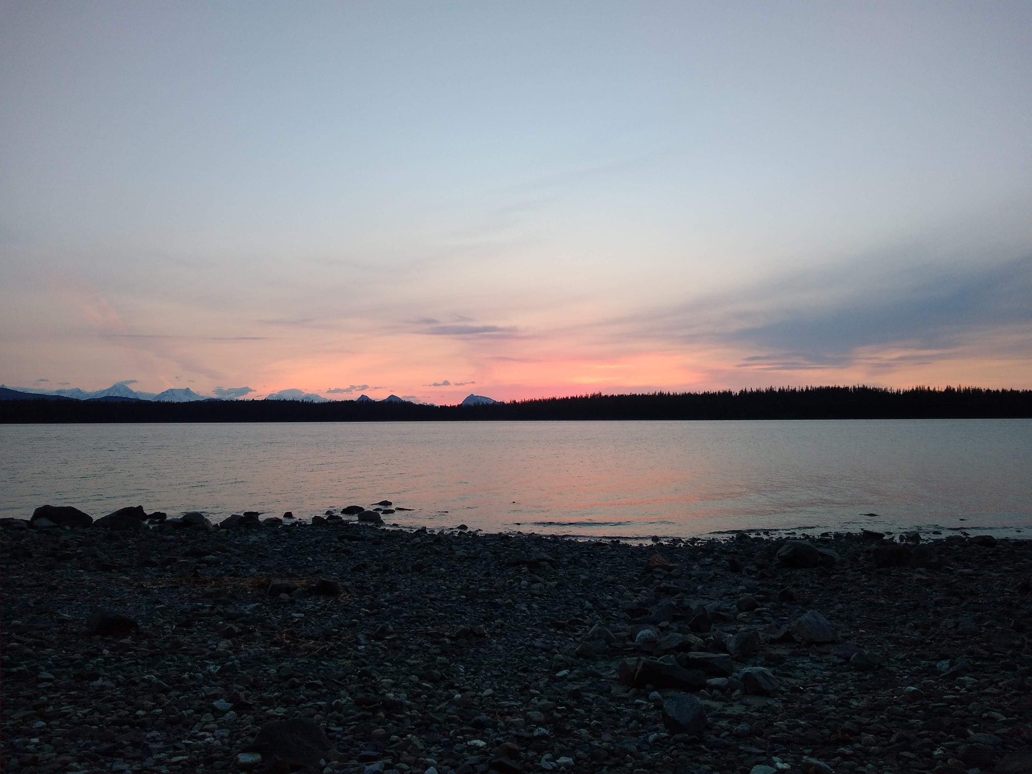 A pink sunset behind distant mountains and a forested island. In the foreground is the beach in the campground at Bartlett Cove campground to visit Glacier bay national park