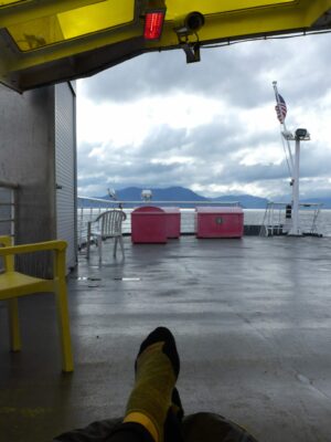 A person's feet are visible in the foreground on the deck of a ship under a cover. you can see the back of the ship, with an American flag, and water and mountains behind on the way to visit Glacier Bay national park