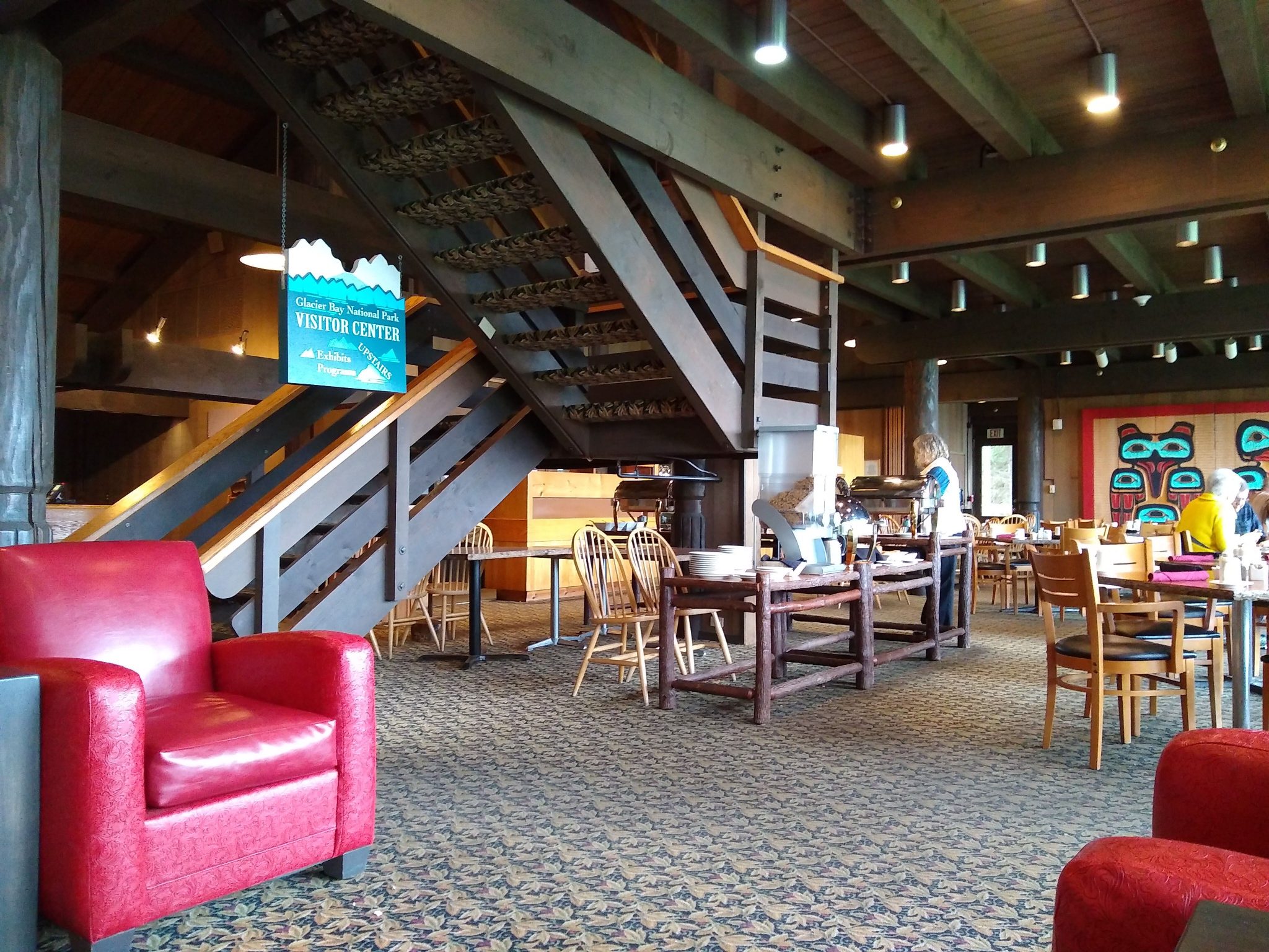 A lodge lobby has a staircase in the middle and red lounge chairs around. There is also a restaurant, with wooden tables and chairs and Tlingit art on the walls. The roof is made of wood and there is a patterned carpet