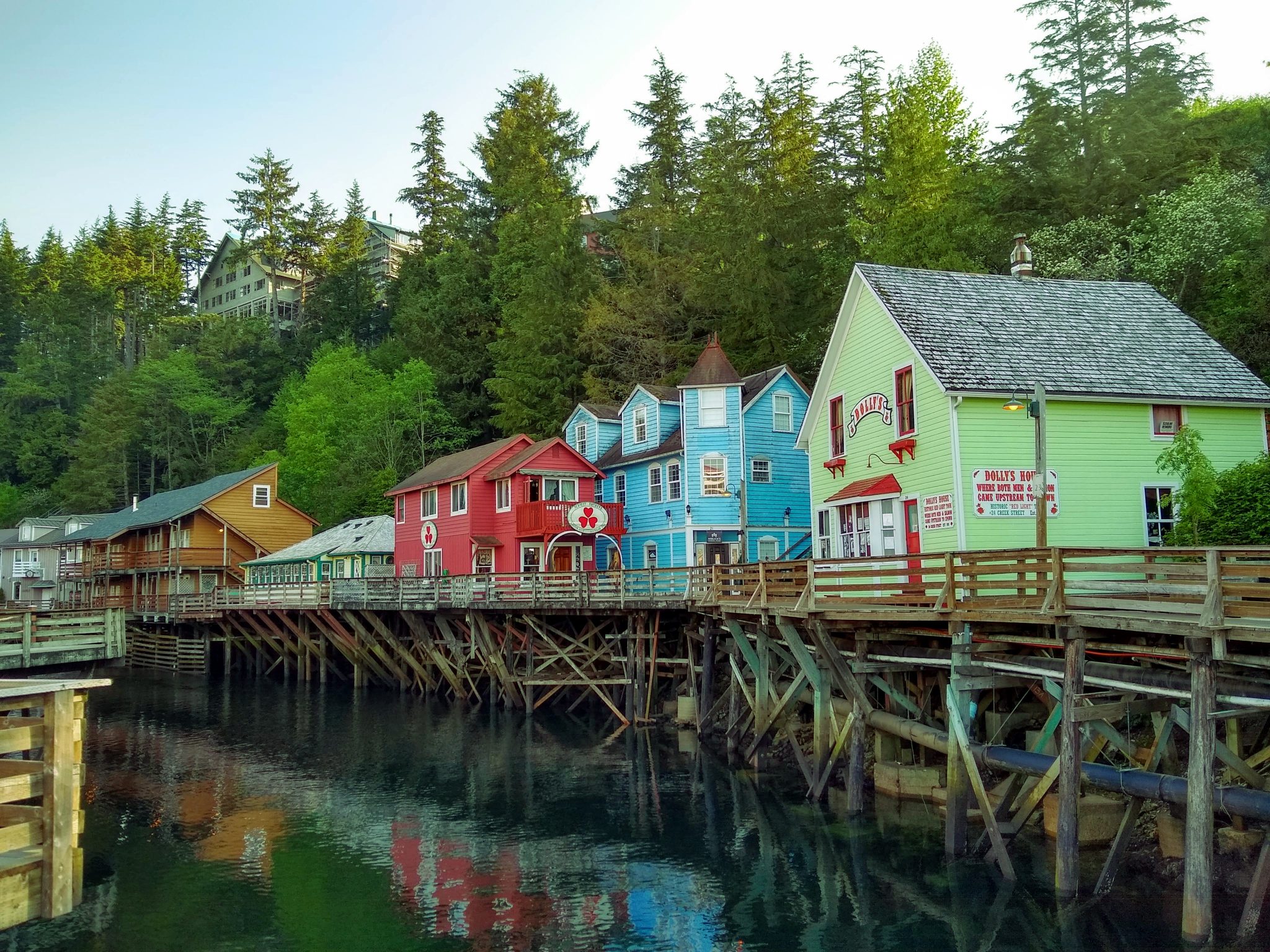 Several brightly colored historic wooden buildings are built on wooden piers above the water. The water is calm and the buildings are reflected in it. There are evergreen trees behind the buildings on the hill