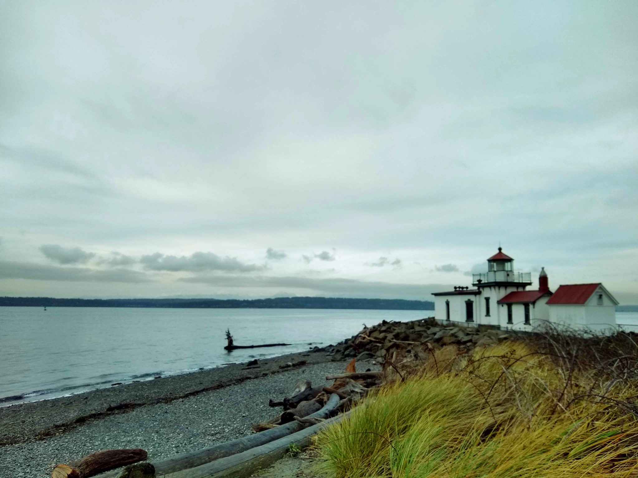 Outdoor activities in Seattle include beaches such as this one by the Westpoint lighthouse. The lighthouse is on a gravel beach with driftwood and rocks. The water is calm and forested hills are seen across the water. It's an overcast day