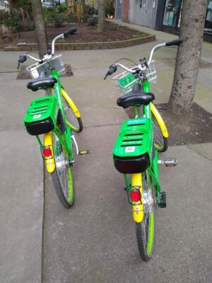 Two green and yellow electric bike share bikes parked next to each other on a Seattle sidewalk. Use a bike share e-bike to bike to alki beach!