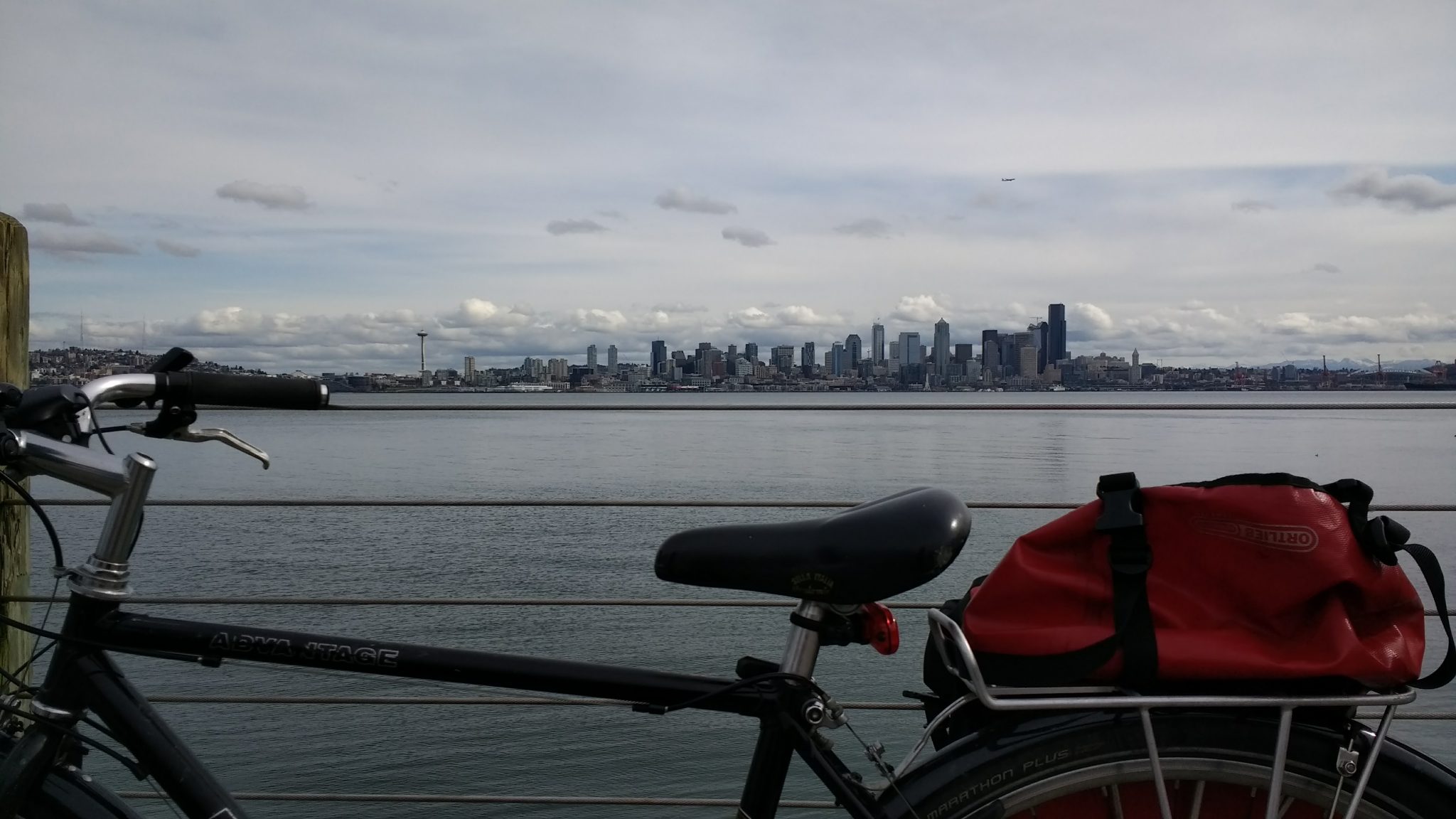 Outdoor activities in Seattle include cycling on the cities many trails. A plain black bike with a red back is in the foreground against a wire railing. There is gray water and gray sky. Across the water the Seattle skyline is visible, including the space needle
