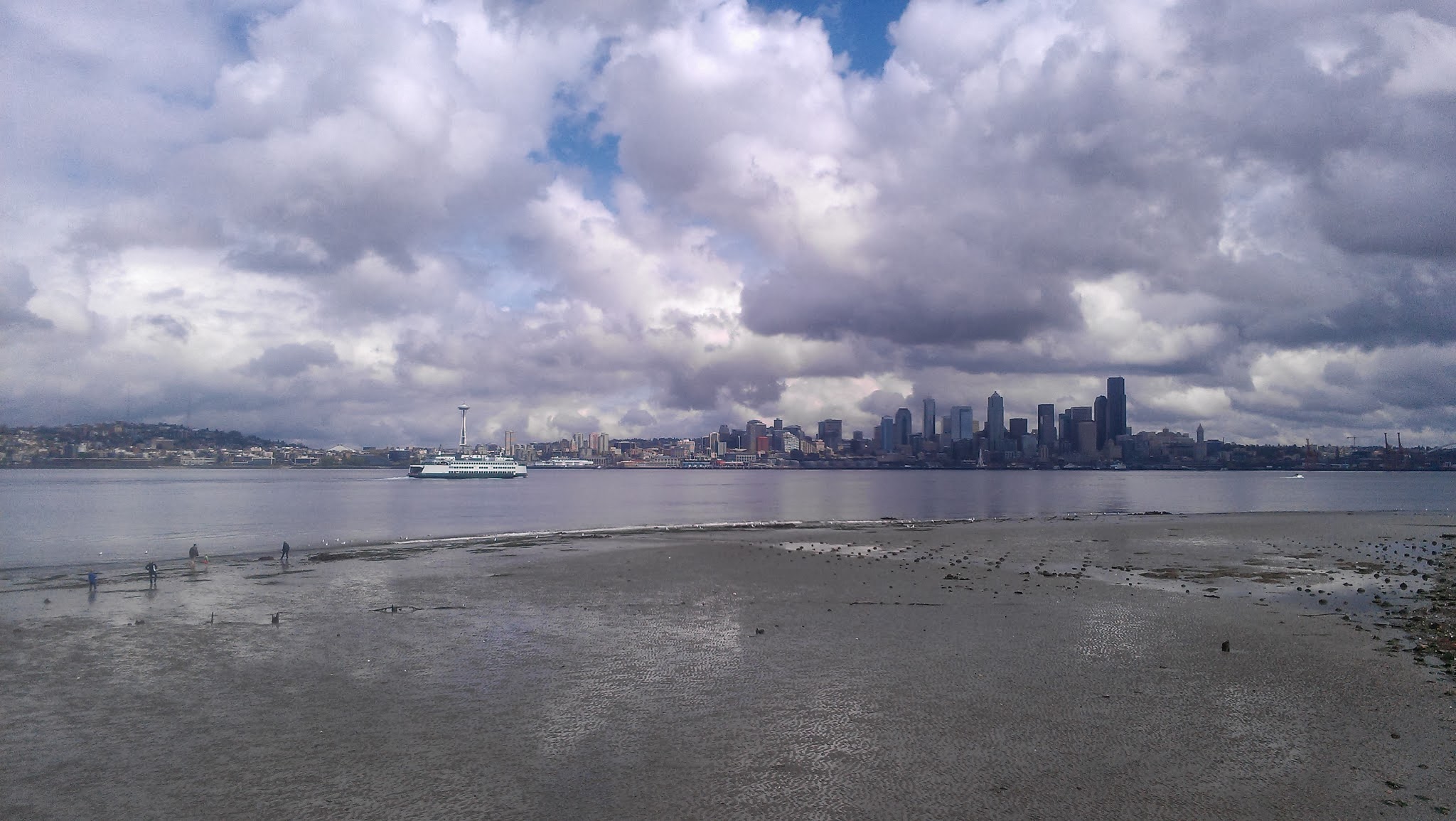A wide tideflat at low tide. There is a bay behind with a ferry arriving in Seattle. The Seattle skyline is visible in the background. There are lots of puffy clouds.